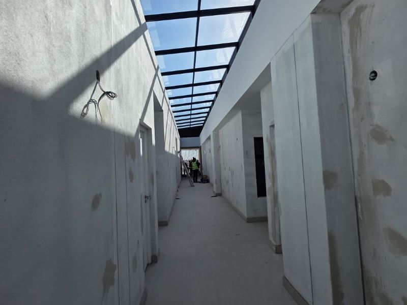 A long, unfinished hallway featuring a large overhead skylight, currently under construction with exposed plaster walls and pending electrical finishes.