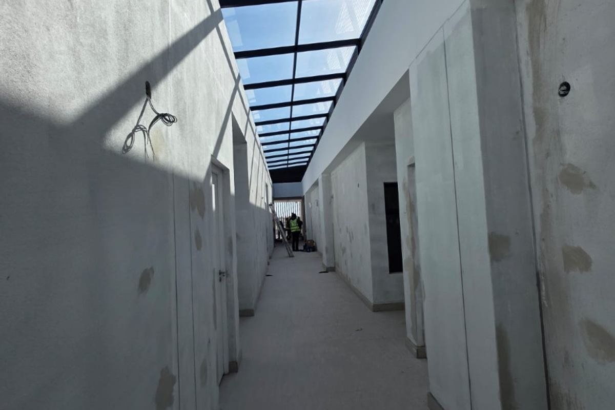 A long, unfinished hallway featuring a large overhead skylight, currently under construction with exposed plaster walls and pending electrical finishes.