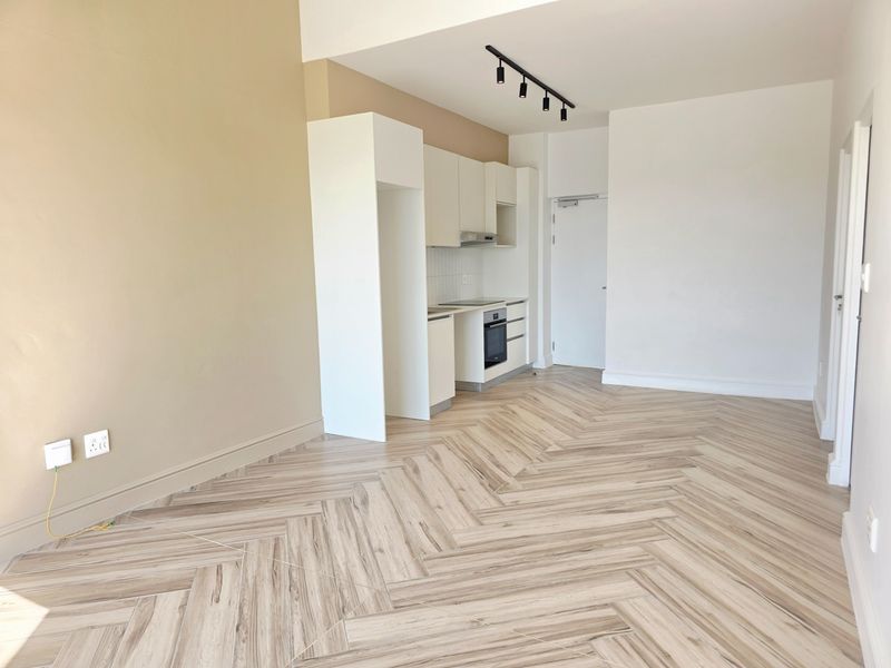 Modern open-plan living area featuring stylish herringbone tile flooring, neutral walls, and a compact, well-appointed kitchen with built-in appliances and track lighting.