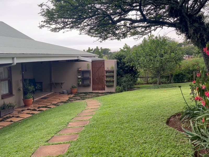 A well-maintained rear garden featuring a stone paver pathway, lush lawn, mature trees, and a covered patio area with a timber gate.