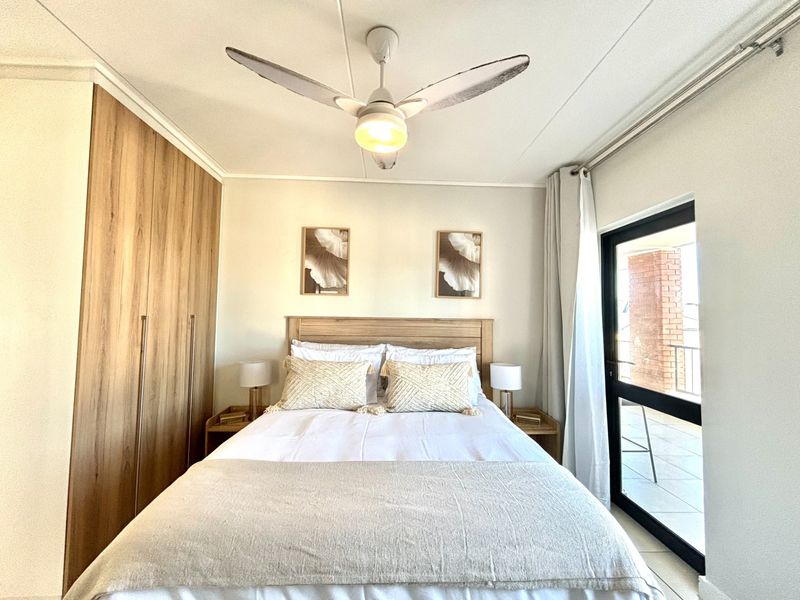 A well-maintained bedroom featuring built-in wooden cupboards, a ceiling fan, and direct access to a private balcony through a sliding glass door.