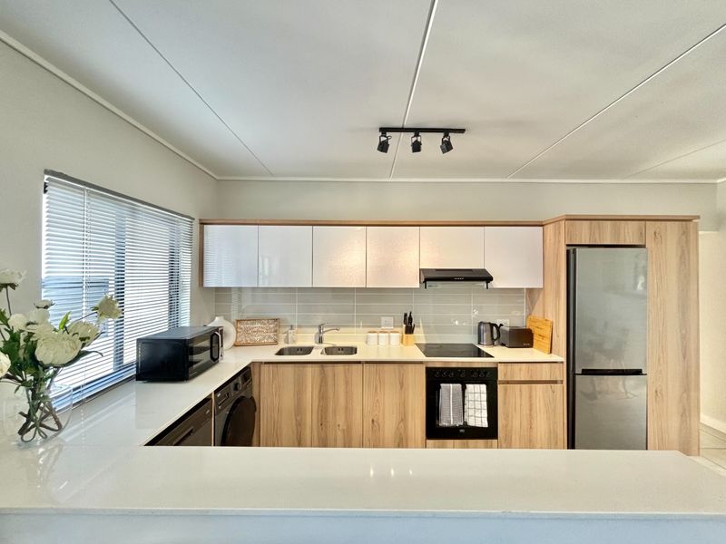 Modern kitchen featuring light wood-grain cabinetry, white stone countertops, tiled backsplash, and integrated appliances with contemporary track lighting.