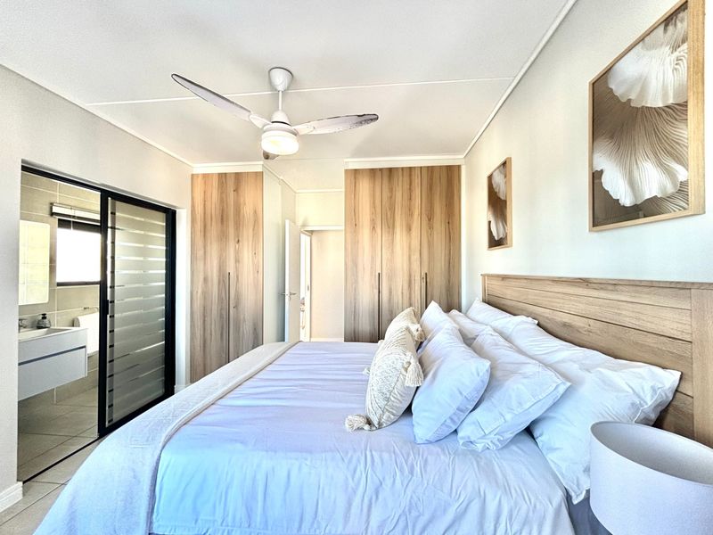 A well-maintained bedroom featuring modern wood-finish built-in cupboards, a ceiling fan, and direct access to an en-suite bathroom through a sliding glass door.