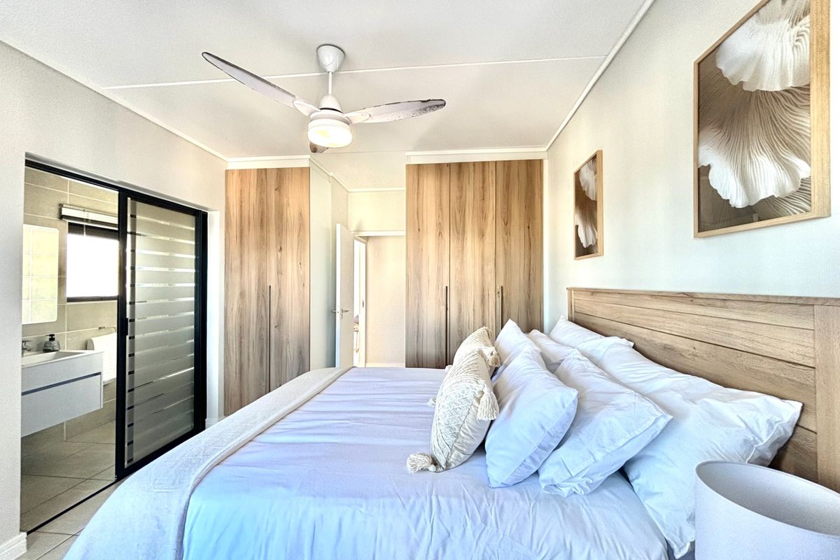 A well-maintained bedroom featuring modern wood-finish built-in cupboards, a ceiling fan, and direct access to an en-suite bathroom through a sliding glass door.