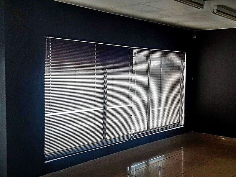 An empty commercial office space featuring dark painted walls, tiled flooring, venetian blinds, and functional overhead fluorescent lighting.