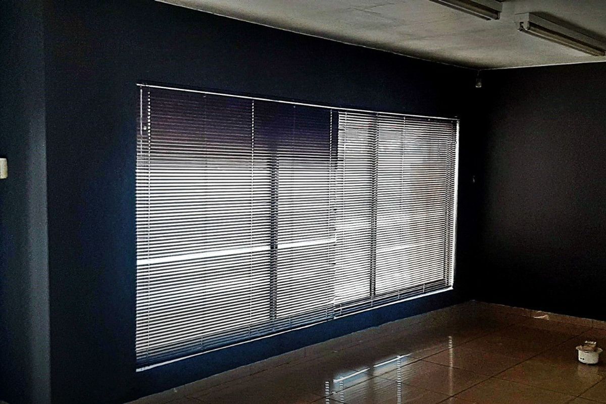 An empty commercial office space featuring dark painted walls, tiled flooring, venetian blinds, and functional overhead fluorescent lighting.