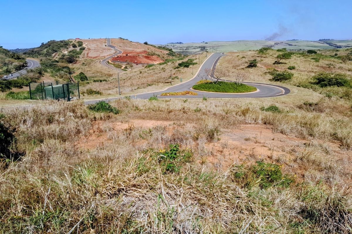 An elevated view of a vacant residential plot overlooking a paved road, roundabout, and surrounding undeveloped landscape.