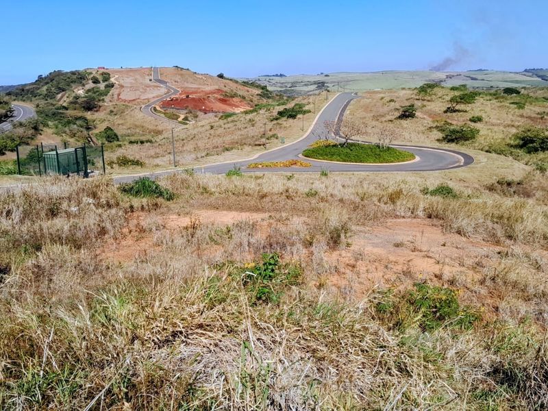 An elevated view of a vacant residential plot overlooking a paved road, roundabout, and surrounding undeveloped landscape.
