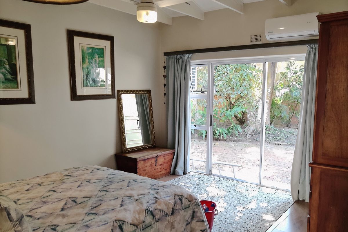 A well-lit bedroom featuring exposed timber ceiling beams, a ceiling fan, split-system air conditioning, and sliding glass doors providing direct access to the garden.