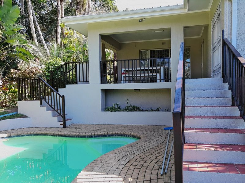 Outdoor pool area featuring a paved deck, covered patio with built-in braai, and concrete stairs with metal railings leading to the main house.