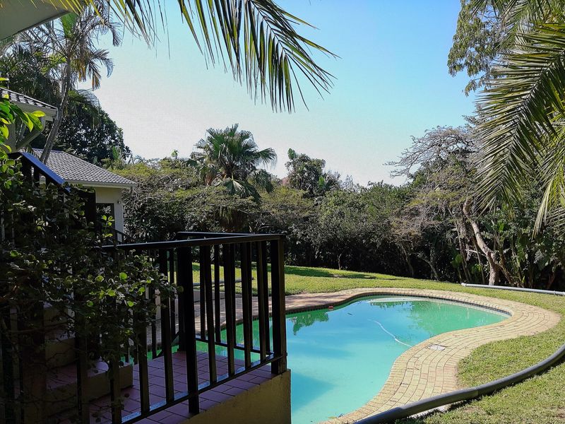A view of a swimming pool with brick coping, set within a landscaped garden area, viewed from an elevated tiled patio.