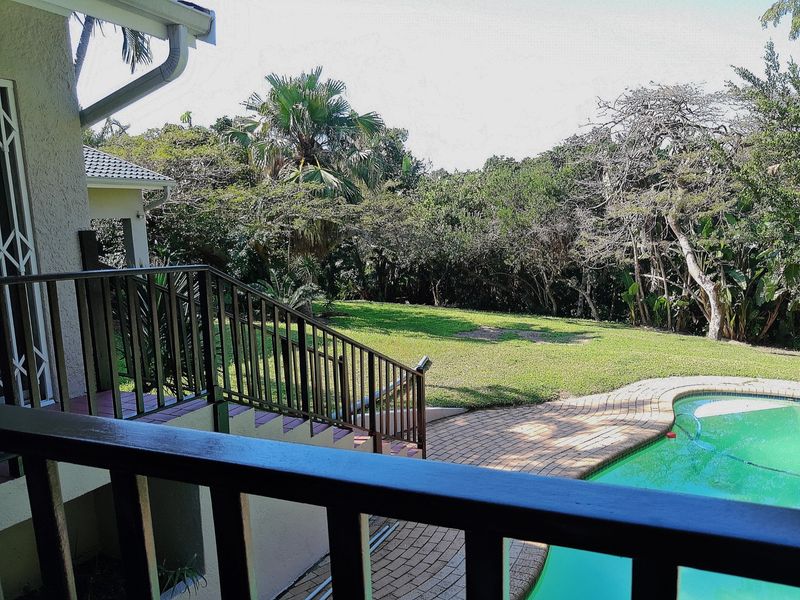 Outdoor view from a balcony overlooking a swimming pool with brick paving and a lush garden setting.