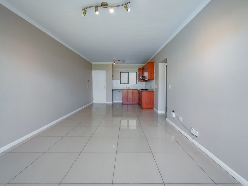 An open-plan living and kitchen area featuring neutral wall tones, large format floor tiles, and a functional kitchen with built-in cabinetry and appliances.