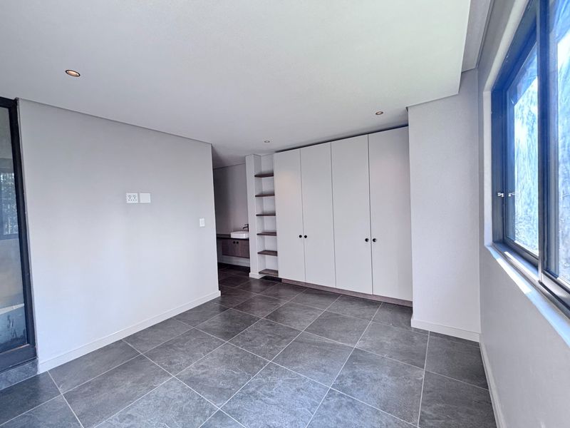 A modern, empty bedroom featuring tiled flooring, built-in cupboards, recessed lighting, and an integrated vanity area with a basin.