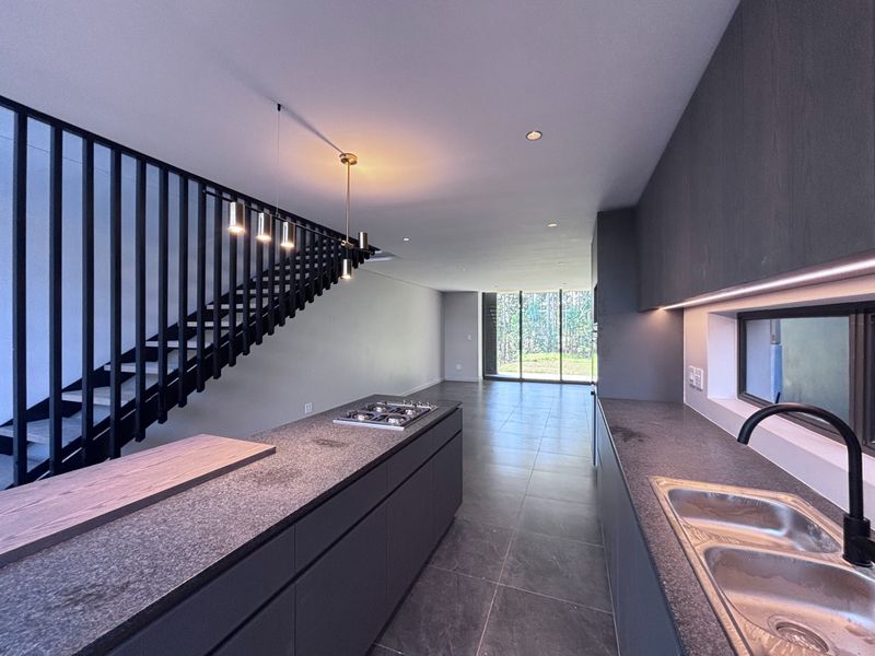 Modern open-plan kitchen and living area featuring sleek dark cabinetry, stone countertops, a gas hob, and a striking staircase with a contemporary metal balustrade.