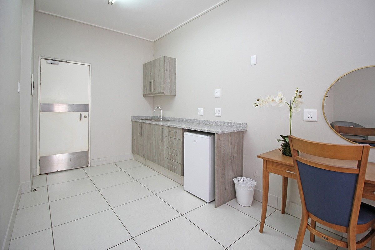 A compact kitchenette featuring light wood-tone cabinetry, stone-look countertops, a stainless steel sink, and durable tiled flooring in a clean, functional retirement unit.