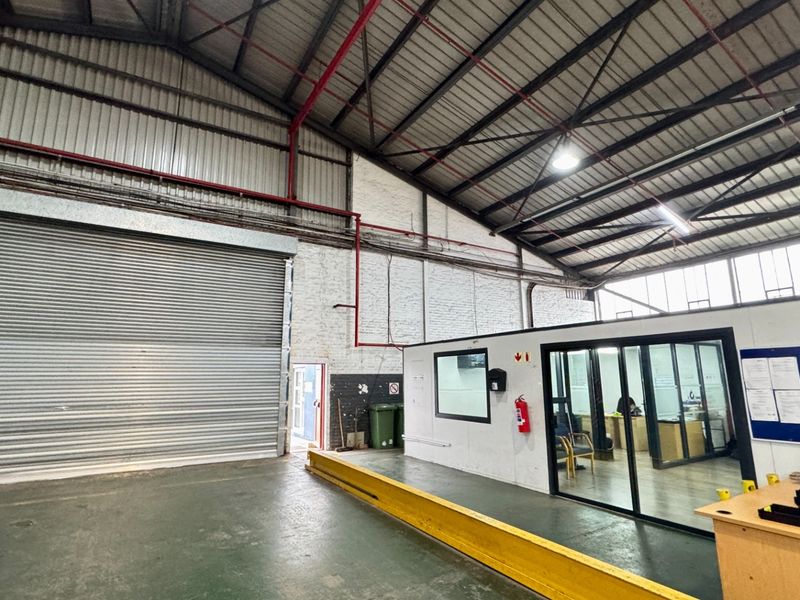 Spacious industrial warehouse area featuring high ceilings with exposed steel trusses, a large roller shutter door, and an integrated partitioned office space.