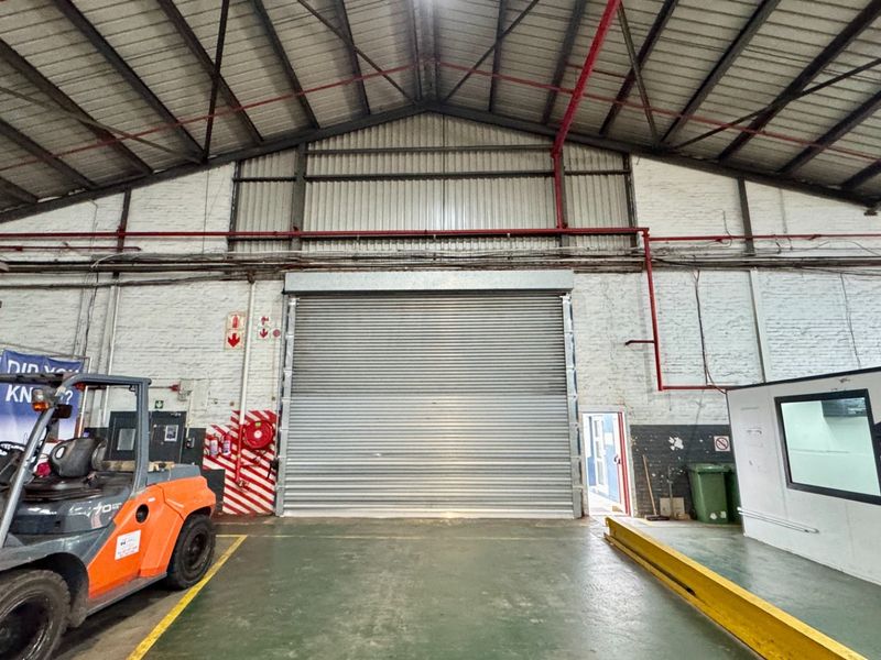 Spacious industrial warehouse area featuring high ceilings with exposed steel trusses, a large roller shutter door, and an integrated office enclosure.