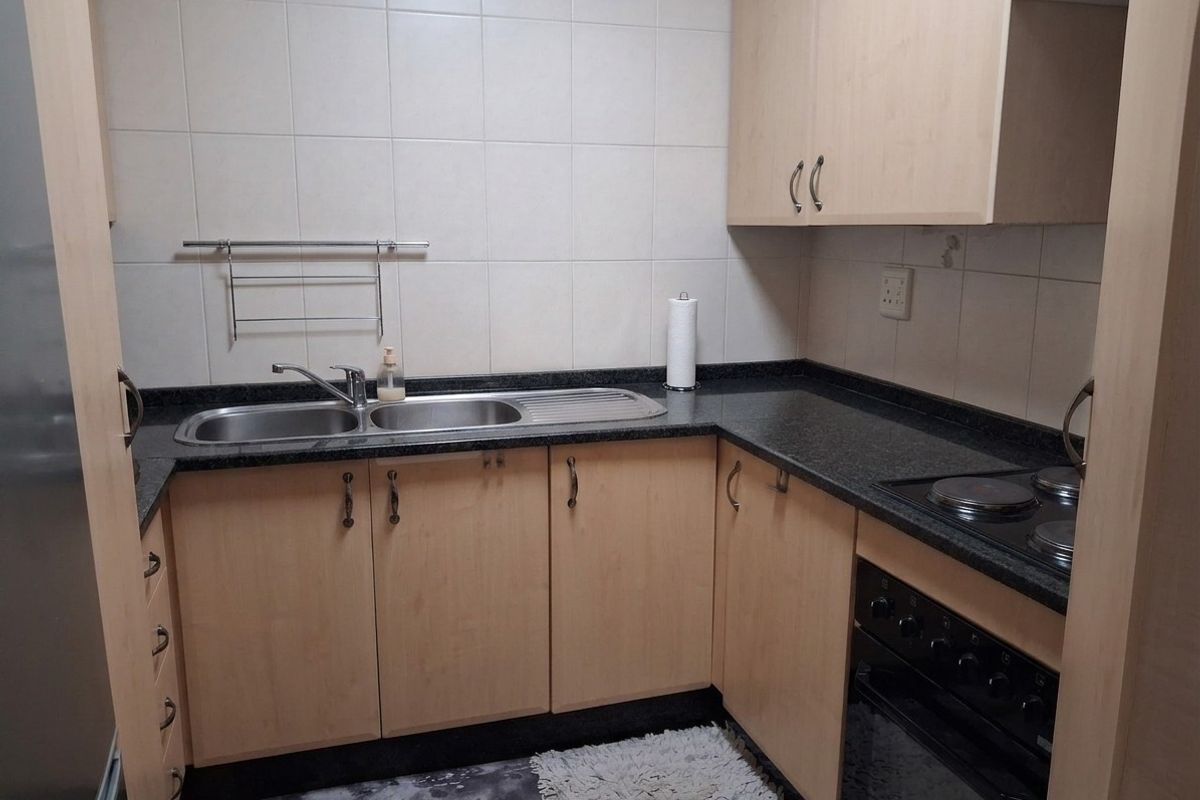 Compact kitchen featuring light wood-finish cabinetry, dark granite countertops, a stainless steel double sink, and a built-in electric oven with hob.