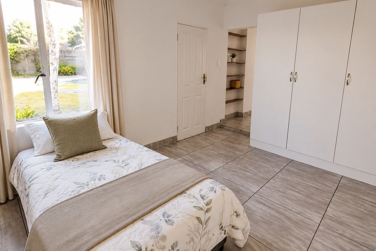 A bright bedroom featuring neutral tiled flooring, built-in cupboards, and built-in shelving, with a large window providing natural light and garden views.
