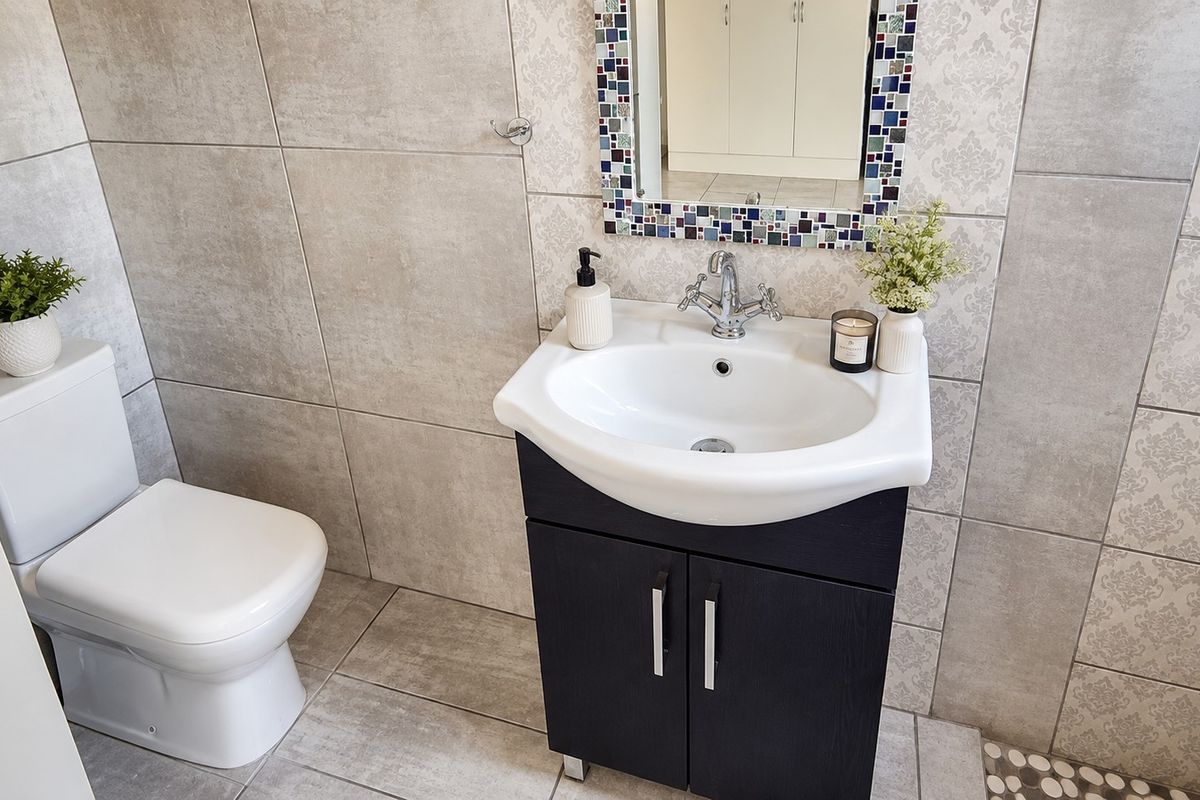A modern bathroom featuring a dark vanity cabinet, ceramic basin with chrome mixer tap, mosaic-framed mirror, and neutral-toned wall and floor tiling.