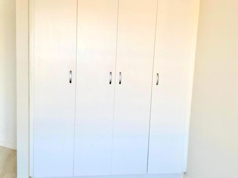 A bright bedroom featuring modern light-toned laminate flooring and a clean, white built-in wardrobe with sleek handles.
