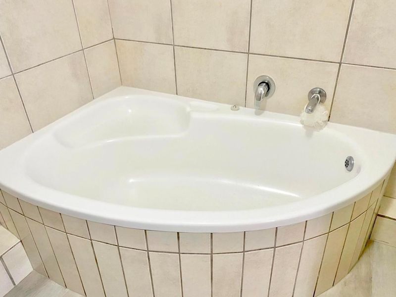 A corner bathtub featuring neutral ceramic wall tiling and wall-mounted chrome fixtures in a clean, functional bathroom space.