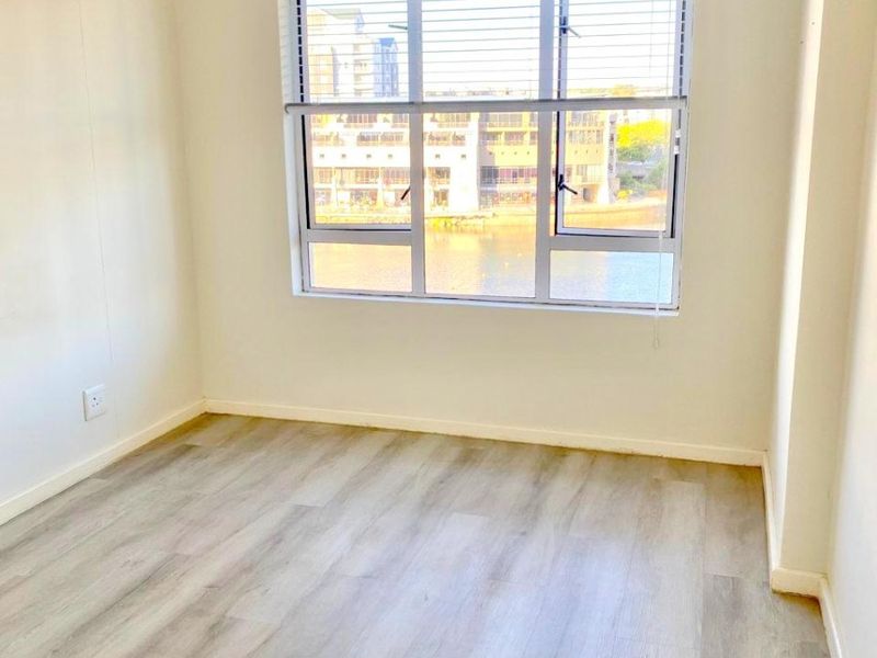 A bright, empty bedroom featuring modern light-toned laminate flooring, neutral walls, and a large window with venetian blinds overlooking the water.