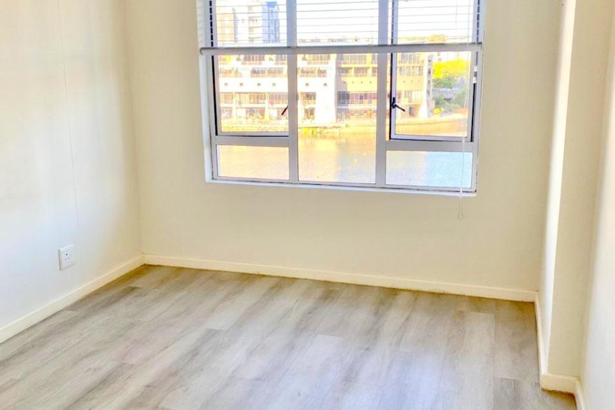 A bright, empty bedroom featuring modern light-toned laminate flooring, neutral walls, and a large window with venetian blinds overlooking the water.