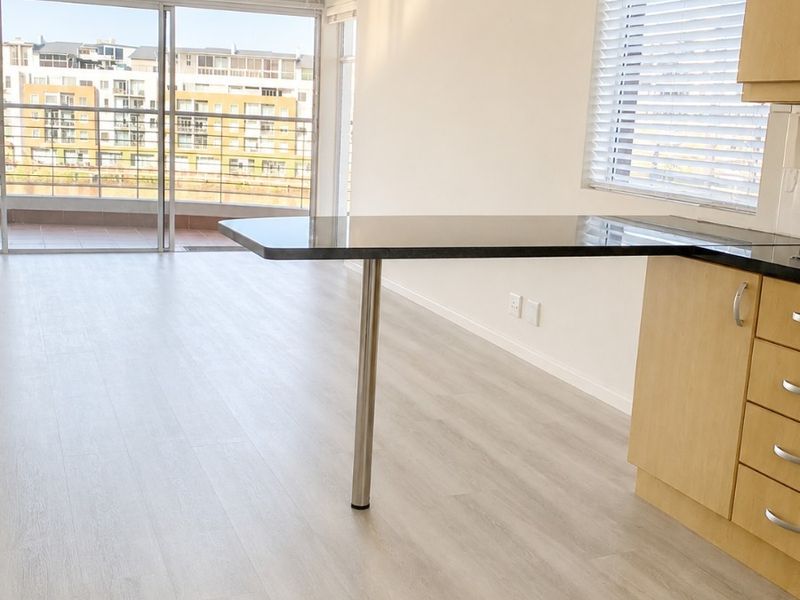 Bright open-plan living area featuring light laminate flooring, built-in kitchen cabinetry with a breakfast bar, and sliding doors leading to a balcony.
