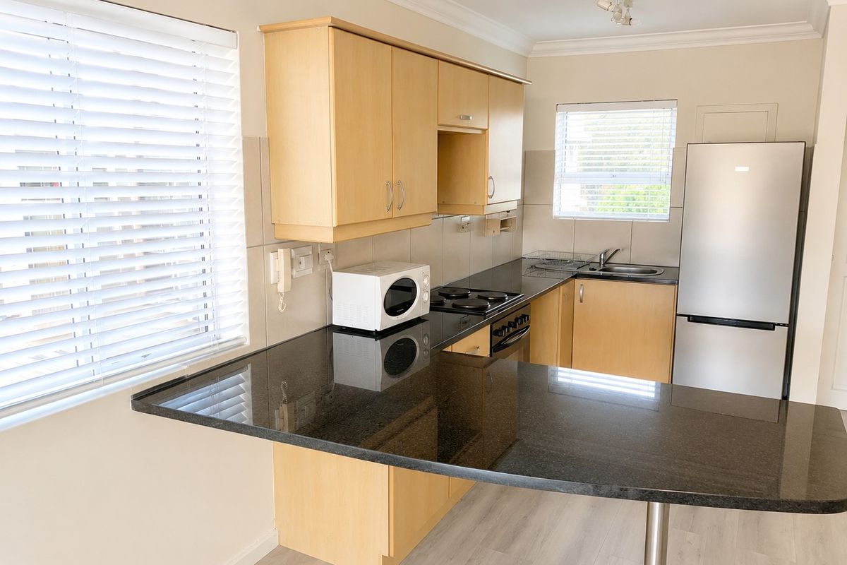 A functional kitchen featuring light wood cabinetry, black granite countertops, an electric hob, and an integrated sink with natural light from dual windows.