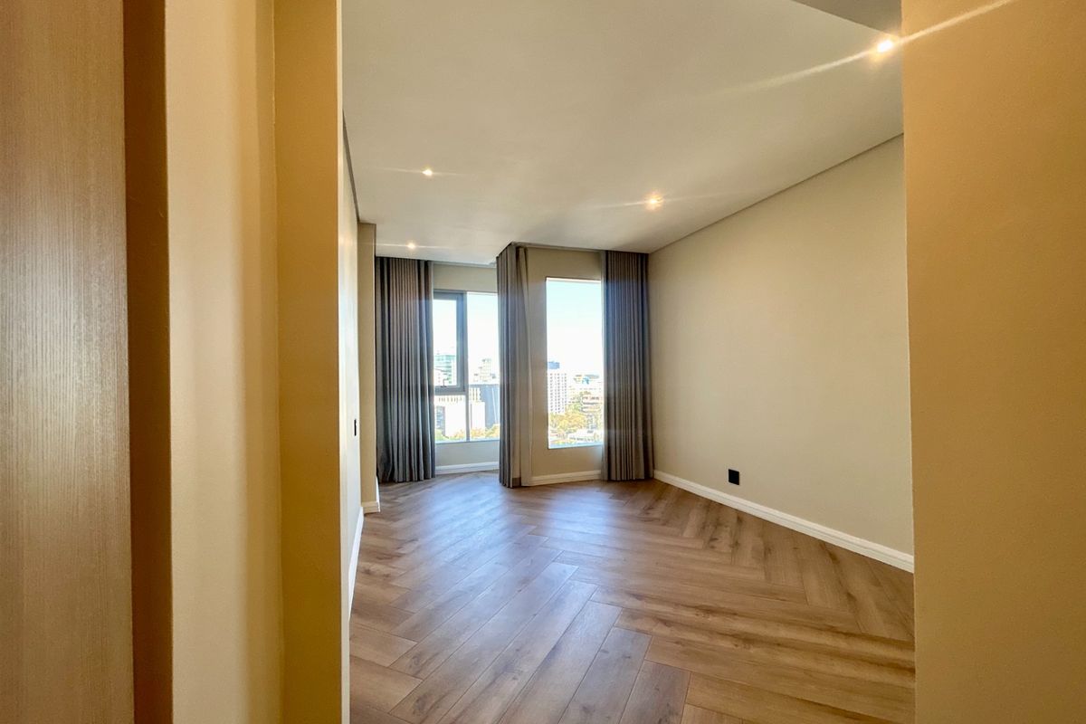 A bright, empty bedroom featuring modern herringbone wood flooring, recessed lighting, and large windows with neutral curtains offering an urban view.
