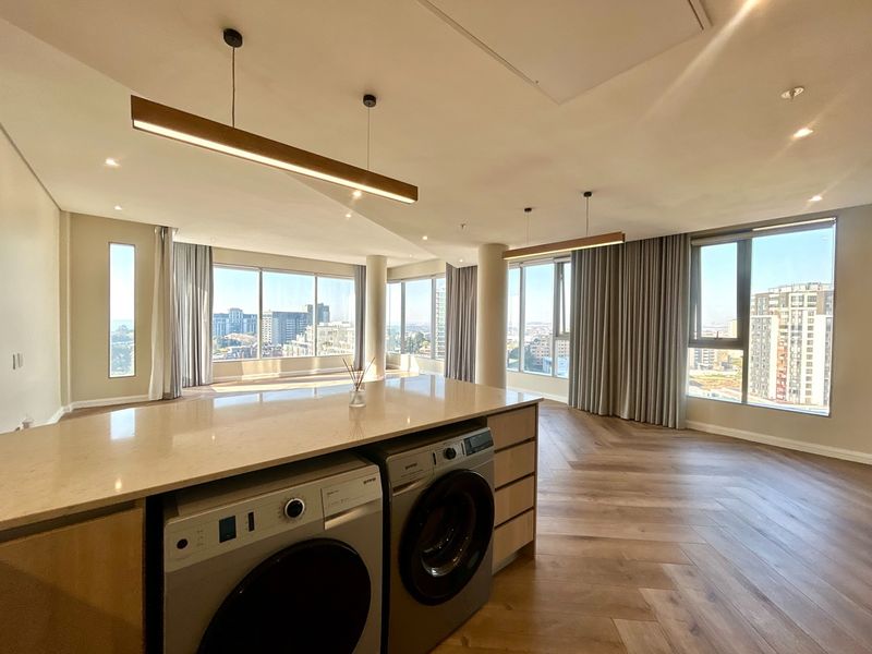 Spacious open-plan living area featuring herringbone timber flooring, floor-to-ceiling windows with city views, and a modern kitchen island with integrated laundry appliances.