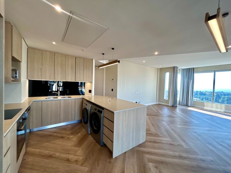 Modern open-plan living area featuring a sleek kitchen with light wood cabinetry, stone countertops, integrated appliances, and elegant herringbone timber flooring with expansive views.