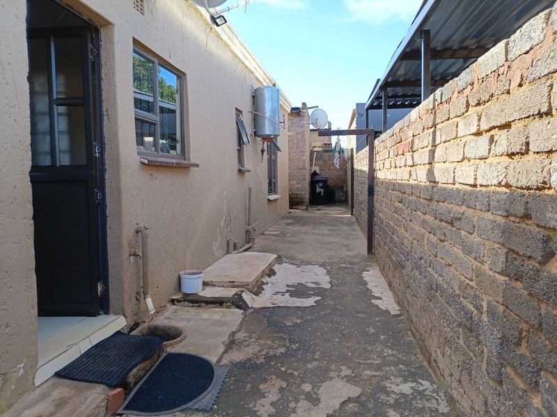 A narrow rear exterior walkway featuring a concrete path, brick boundary wall, and external geyser unit.