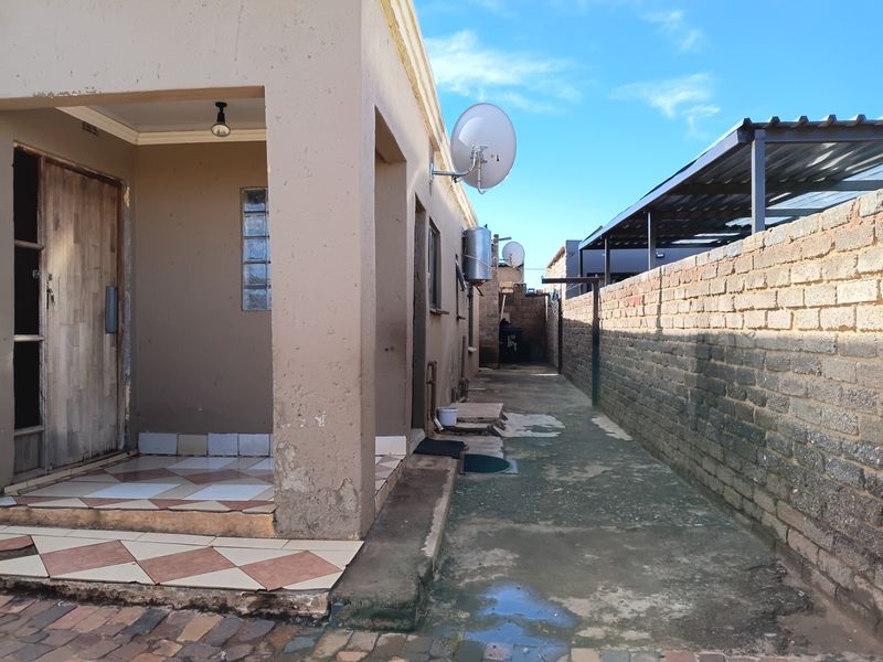 Rear exterior view showing a tiled patio area, brick boundary wall, carport structure, and wall-mounted geyser unit.