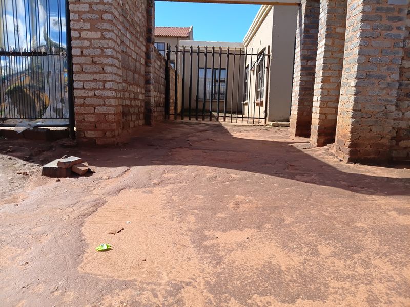 Front entrance area featuring brick pillars and a metal security gate, leading to an unpaved driveway and the main building facade.