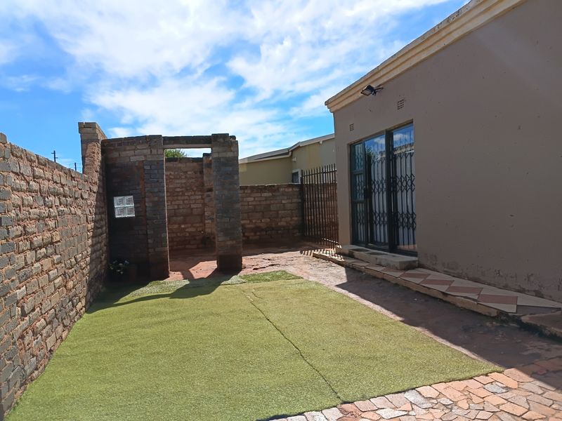 Rear exterior view featuring a paved patio area, artificial lawn, and a brick boundary wall with an unfinished outdoor structure.