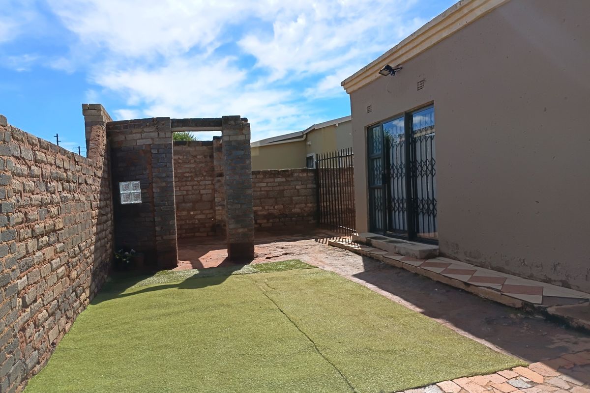 Rear exterior view featuring a paved patio area, artificial lawn, and a brick boundary wall with an unfinished outdoor structure.