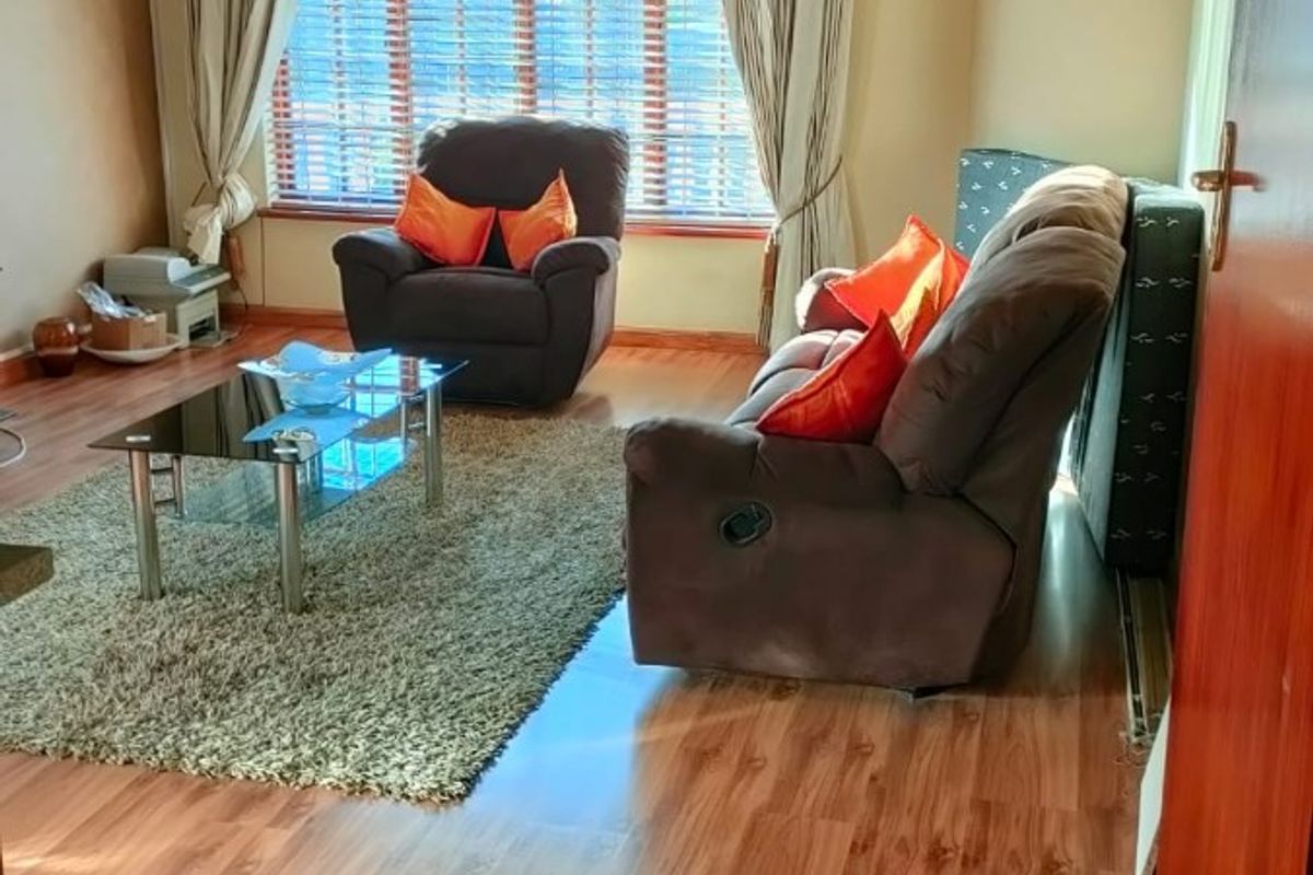 A comfortable living room featuring warm-toned laminate flooring, a decorative dropped ceiling with recessed lighting, and large windows fitted with venetian blinds.