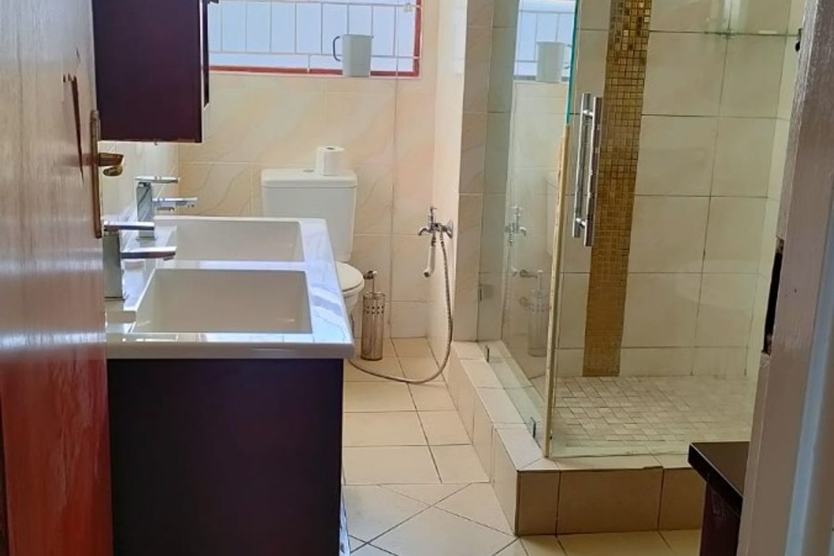 A functional bathroom featuring a double vanity, a glass-enclosed shower with decorative mosaic tile accents, and neutral floor tiling.