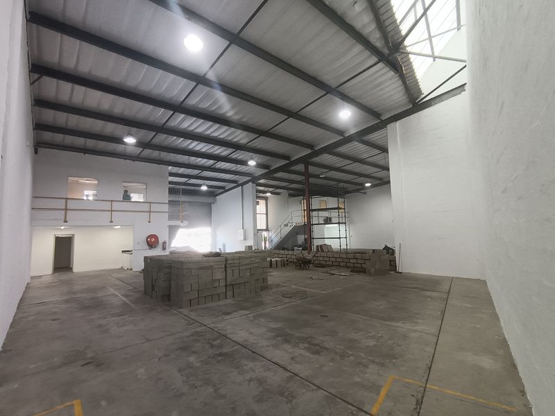 Spacious industrial warehouse unit featuring high ceilings with exposed steel trusses, concrete flooring, a mezzanine level, and a large roller shutter door.