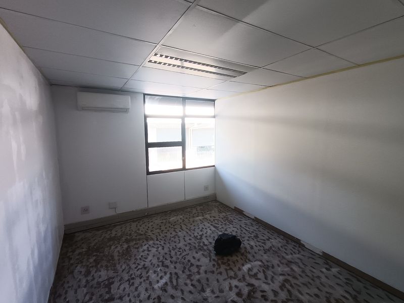 An empty office space featuring a suspended ceiling, recessed lighting, a wall-mounted air conditioning unit, and large windows with aluminium frames.