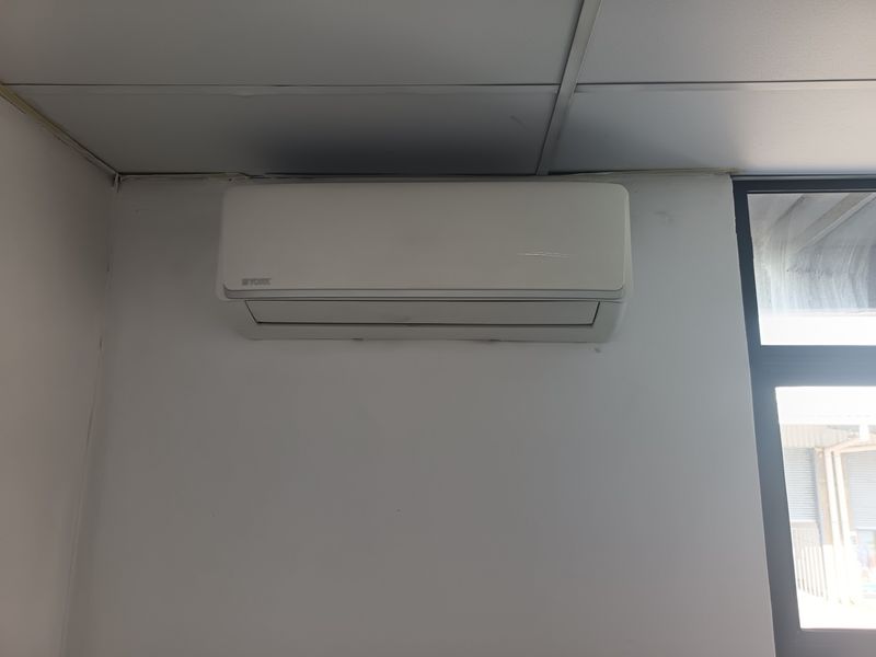 A functional office space featuring a wall-mounted air conditioning unit, suspended ceiling tiles, and a large window with an aluminium frame.