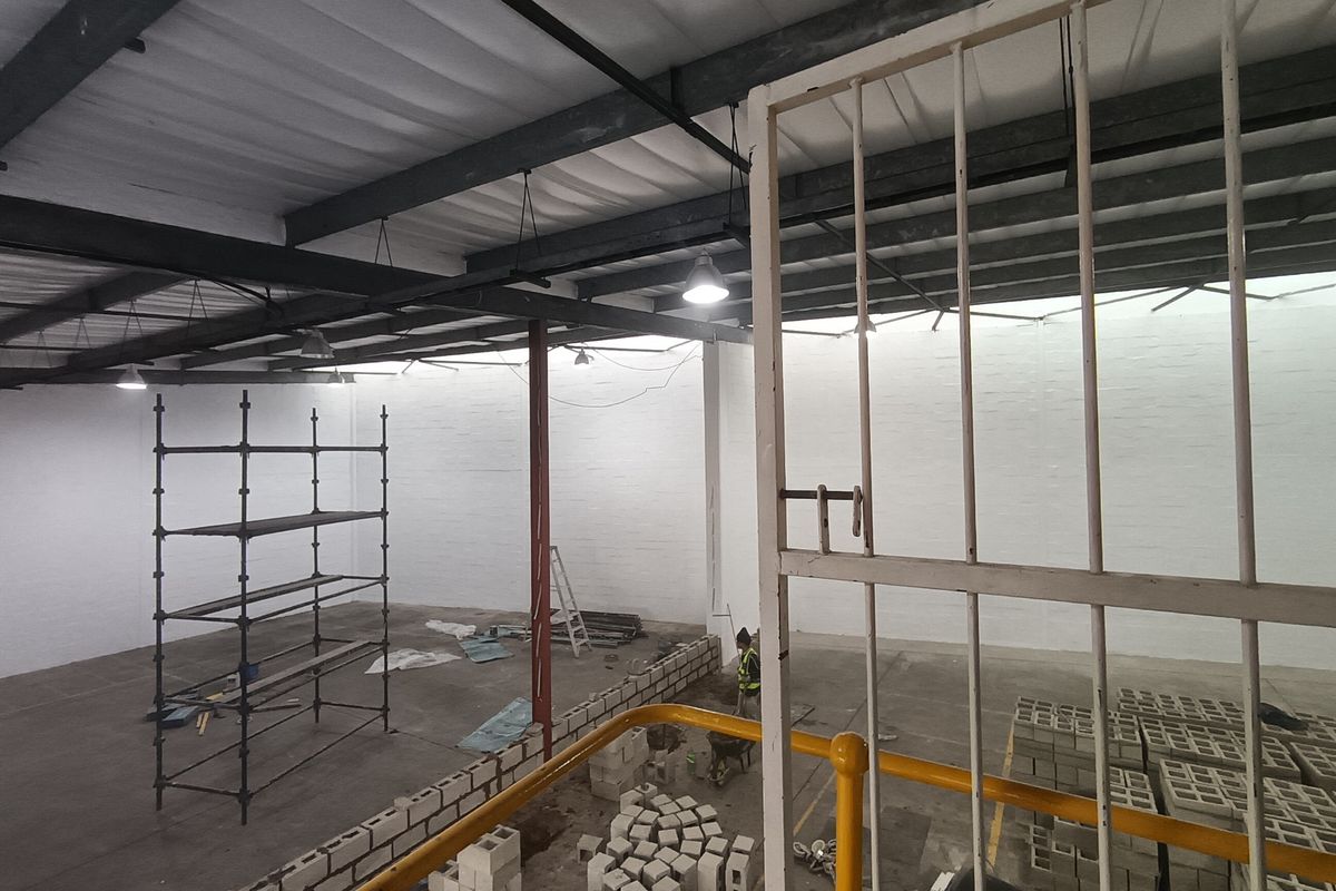Spacious industrial warehouse interior featuring high exposed steel ceilings, painted brick walls, and concrete flooring, currently undergoing renovation.