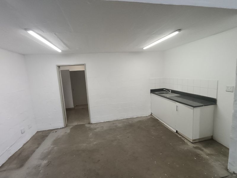 A functional, empty kitchenette area featuring a built-in sink unit, tiled splashback, and industrial-style fluorescent lighting, suitable for office or utility use.