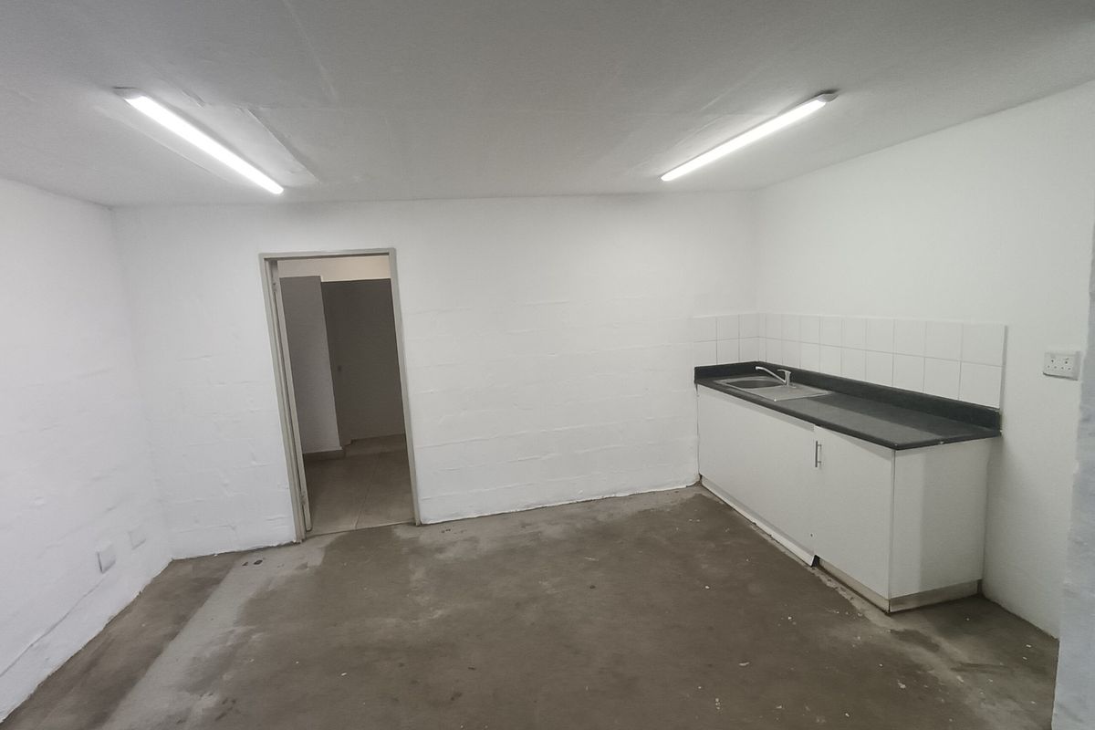 A functional, empty kitchenette area featuring a built-in sink unit, tiled splashback, and industrial-style fluorescent lighting, suitable for office or utility use.