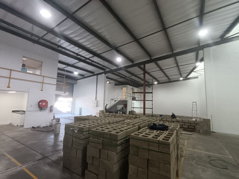 Spacious industrial warehouse unit featuring high ceilings with exposed steel trusses, concrete flooring, a mezzanine level, and a large roller shutter door.