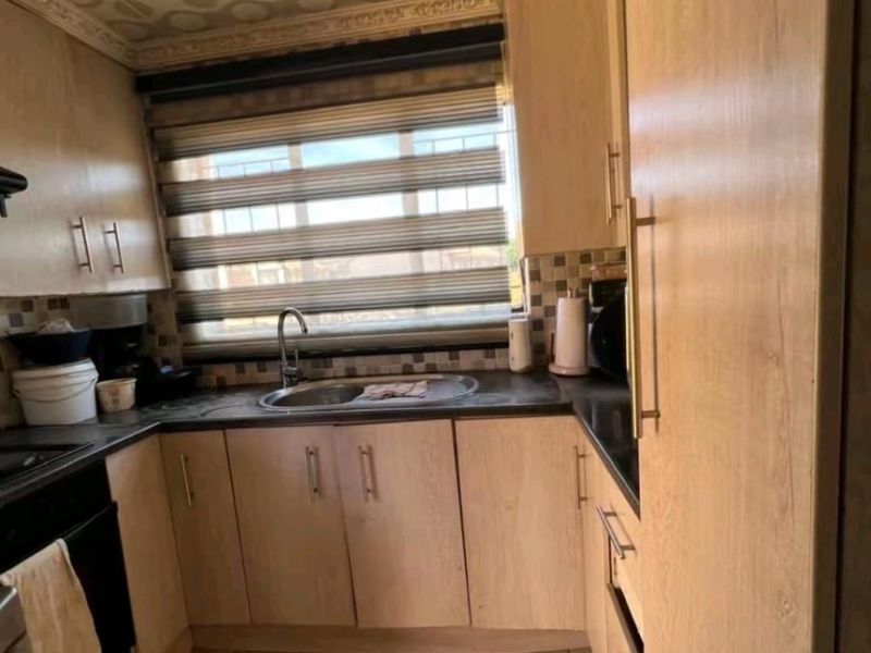 A compact kitchen featuring light wood-finish cabinetry, dark laminate countertops, a stainless steel sink, and tiled flooring with a distinctive patterned ceiling.