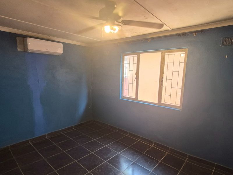 An empty bedroom featuring tiled flooring, a ceiling fan, and a wall-mounted air conditioning unit, with security bars fitted to the window.